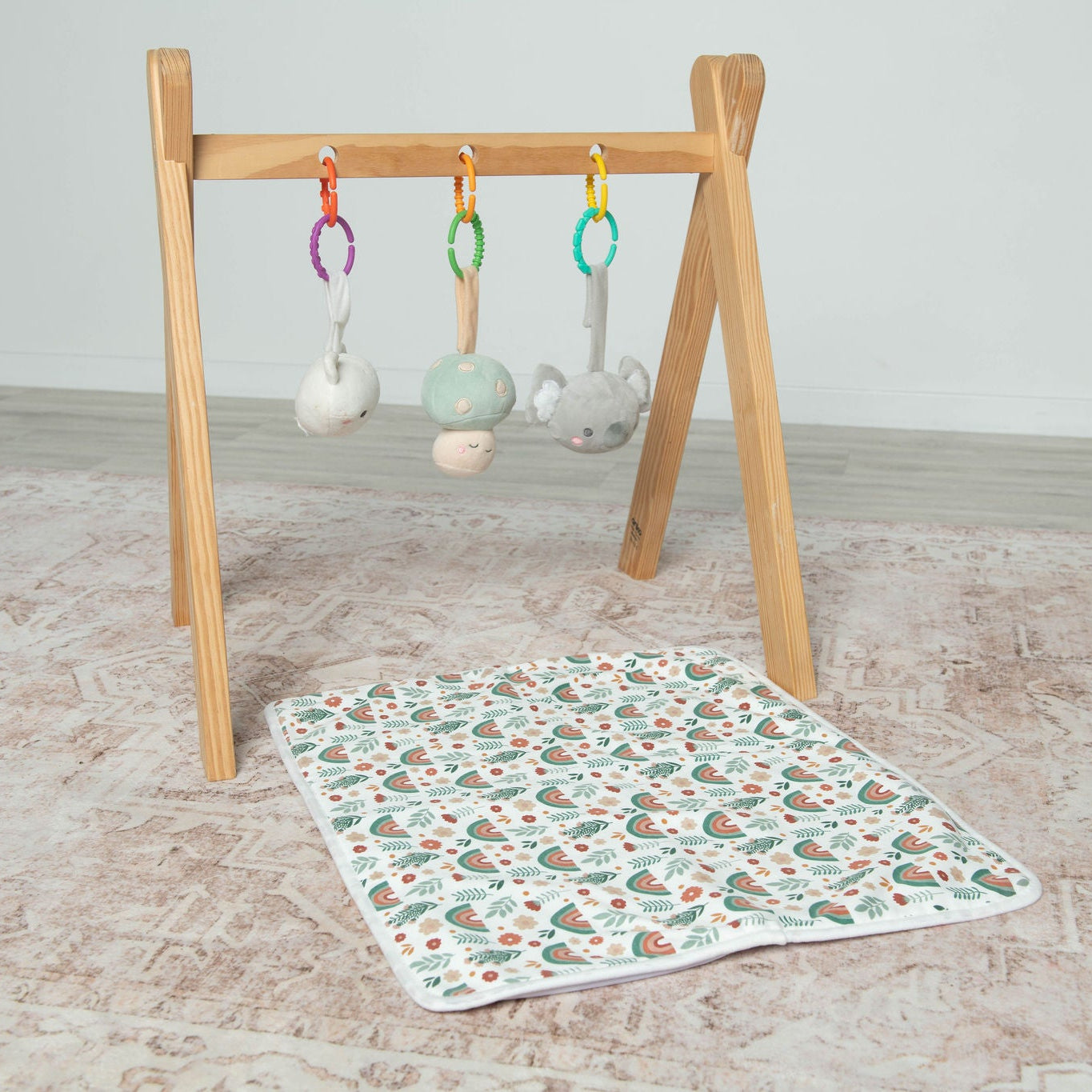 Wooden baby gym with hanging toys and a patterned Spewy Mat on a light wooden floor.